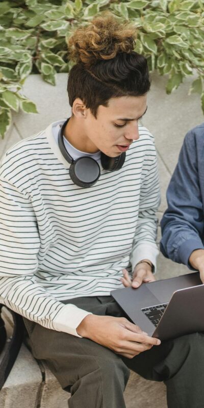 Serious young multiethnic men students in casual clothes studying on laptop while sitting on stone bench in street in daytime with backpack near green plants